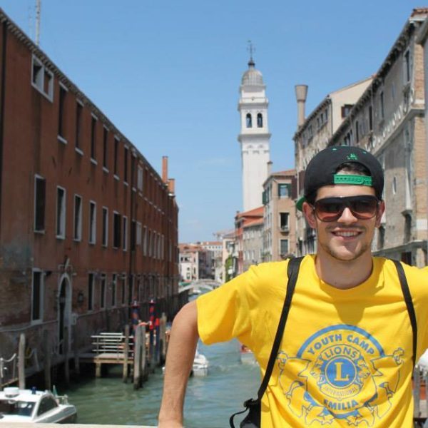 Na foto Augusto sorri, ao fundo passa o rio Veneza, ele está com uma camiseta do LIONS.