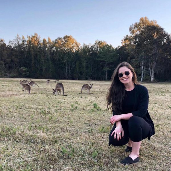 Mulher agachada vestida de preto em um gramado de outono, ao fundo tem cangurus australianos.